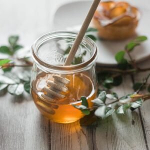 Glass jar filled with honey, featuring a wooden dipper, surrounded by greenery.